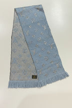 A light blue woven scarf with silver-tone monogram motifs and fringe ends, laid flat on a white surface.