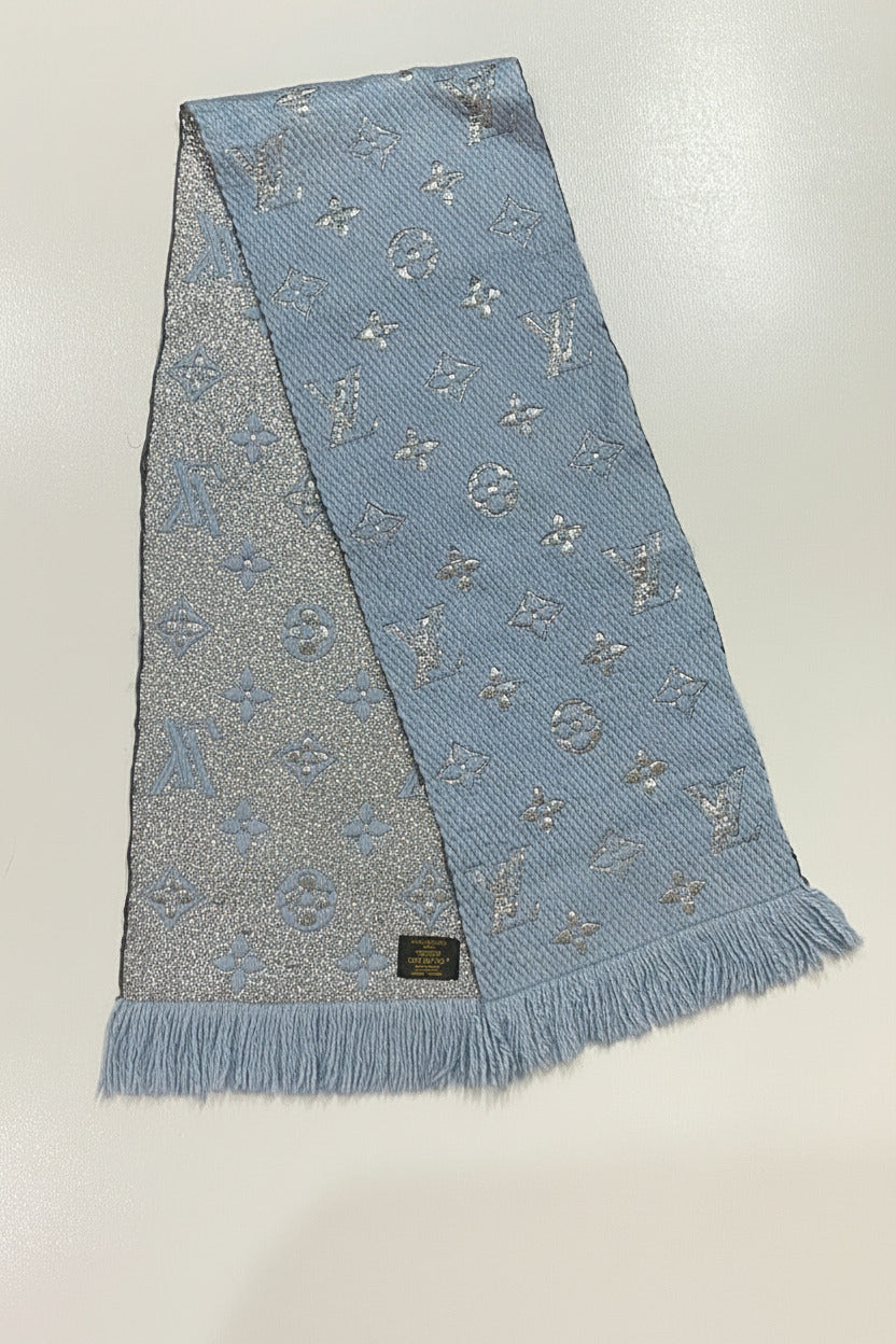 A light blue woven scarf with silver-tone monogram motifs and fringe ends, laid flat on a white surface.