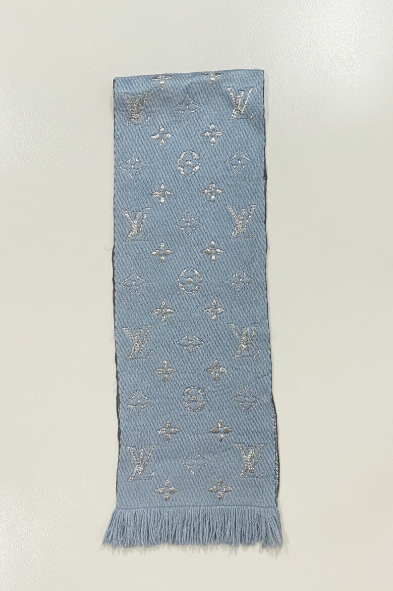 A light blue woven scarf with silver-tone embroidered monogram motifs and fringe at the bottom, laid flat on a white surface.