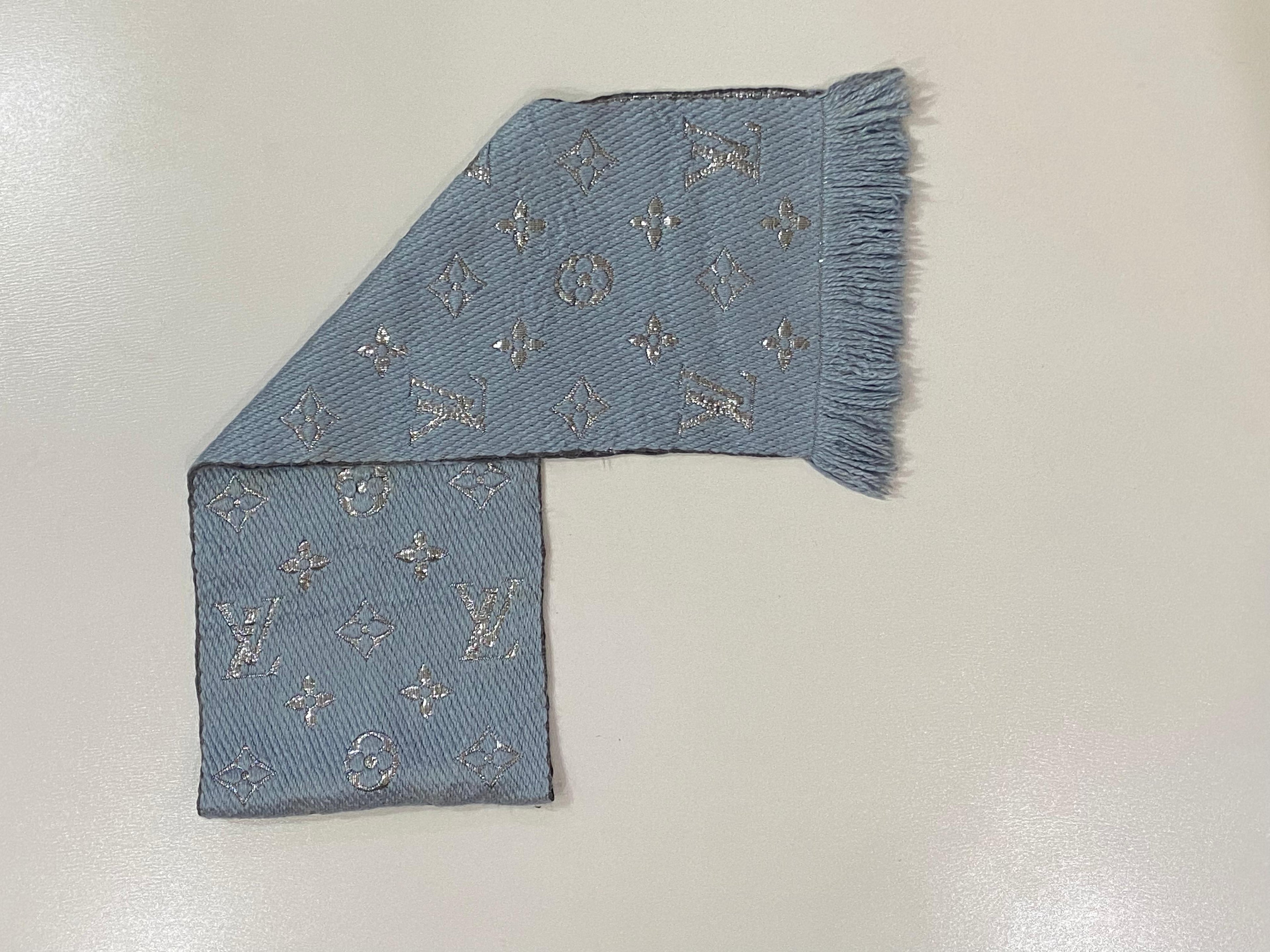 A light blue woven scarf with silver-tone embroidered monogram motifs and fringe trim, laid flat on a light gray surface.
