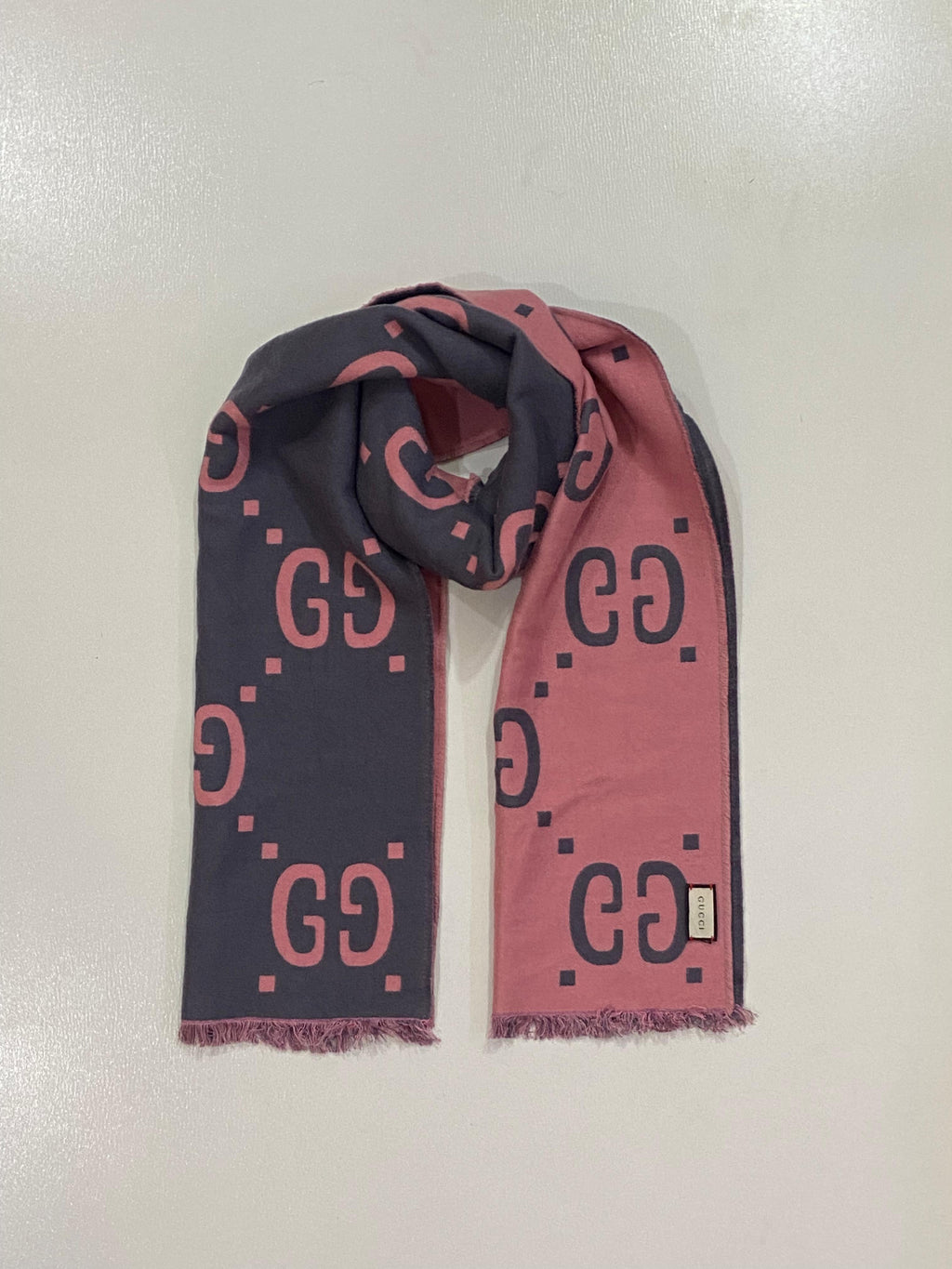 A Gucci scarf with a pink and navy blue color-blocked design featuring the brand's interlocking GG logo pattern, fringed ends, and a small white brand tag.